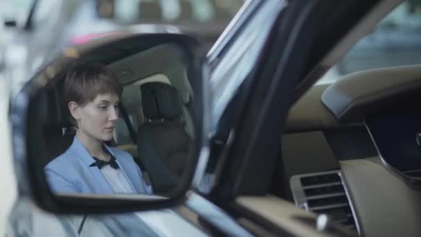 Hermosa joven con un corte de pelo corto examina nuevo auto mientras está sentado en el compartimiento de pasajeros del coche nuevo en la sala de exposición del coche. Mujer bonita exitosa examina nuevo auto. concesionario de coches . — Vídeos de Stock