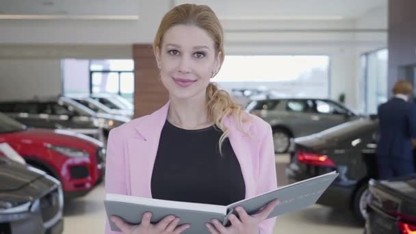 Porträt einer sympathischen Frau in rosa Jacke, die in Großaufnahme auf die Kamera zugeht. Fahrzeuge stehen im Hintergrund. Bild eines lächelnden Verkäufers im Autohaus — Stockvideo