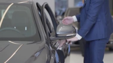 Arabanın içinde modern Bayilik seçerek iş adamı. Uzun boylu adam kapıyı açar ve Otomobil Oto showroom oturur kapatın. Arka planda büyük araba yakın duran bulanık kadın figürleri