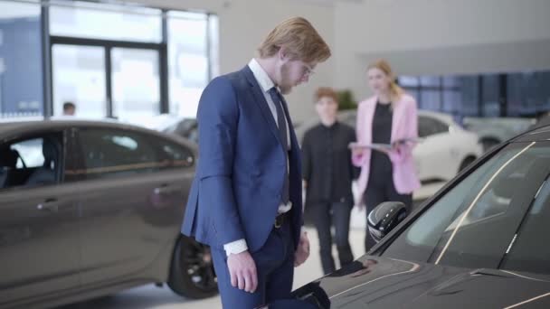 Бізнесмен вибирає машину в дилерському центрі крупним планом. Високий чоловік стоїть біля автомобіля в автосалоні, дивлячись на автомобіль і вивчаючи автомобіль, менеджер показує автомобілі молодій жінці на задньому плані — стокове відео