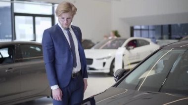 Sakallı işadamı Bayilik arabaya yakın çekim seçme. Siyah arabaya arıyorsunuz ve araç inceleyerek uzun boylu adam. Bulanık insanlar arka planda yürümek. Otomobil Fuarı. Araç Edilmesidir.