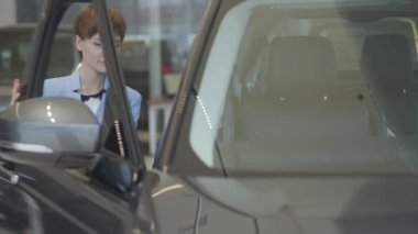 Genç kadın mavi takım arabanın kapısını açar ve içine motor gösterisinde oturur formal giyim güzel. Modern kız otomobil satın almak seçerek, titizlikle inceleniyor. Otomatik araç iş kavramı