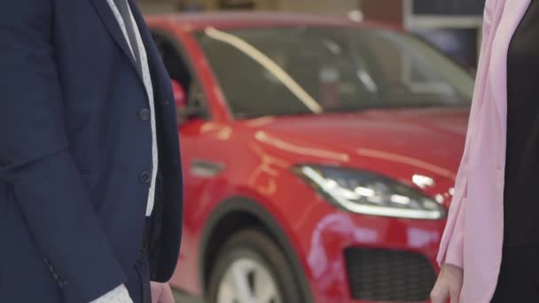 Homme méconnaissable en costume donne les clés à la femme en veste rose de près. Voiture rouge en arrière-plan. Vendeur de voiture remettant les clés de voiture à dame dans le showroom. Concept d'achat d'un véhicule, entreprise automobile — Video