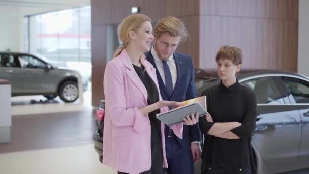 Ευχάριστη γυναίκα σε ροζ σακάκι δείχνει πληροφορίες στο βιβλίο στους πελάτες. Επαγγελματική πωλήτρια βοηθά άνδρας και γυναίκα, για να επιλέξετε το όχημα. Έννοια της αγοράς αυτοκινήτων, auto επιχειρηματικές — Αρχείο Βίντεο