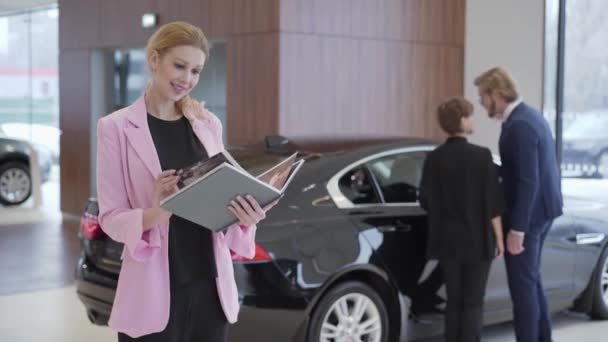 Πορτραίτο του ευχάριστη γυναίκα σε ροζ σακάκι με ένα μεγάλο βιβλίο για τα αυτοκίνητα μπροστά από το ζευγάρι επιλέγοντας το όχημα. Άντρας ανοίγει την πόρτα και η γυναίκα κάθεται στο εσωτερικό. Έννοια της αγοράς αυτοκινήτων, auto επιχειρηματικές — Αρχείο Βίντεο