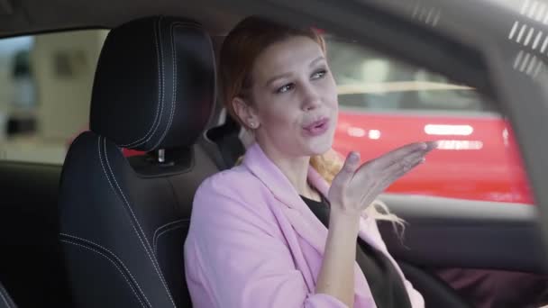 Mujer sonriente confiada en chaqueta rosa en el coche, mirando en el espejo retrovisor y enviando un beso de aire a su reflejo de cerca. Mujer moderna que elige el automóvil en la concesionaria. Concepto de compra de vehículos — Vídeo de stock