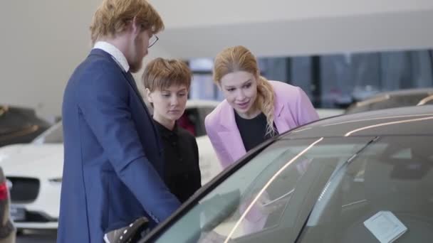 Vendedora de jaqueta rosa discutir o carro com os clientes que estão perto do automóvel e olhando para dentro através da janela. Empresário e sua esposa escolhendo veículo. Motoshow. Sala de exposições do carro . — Vídeo de Stock