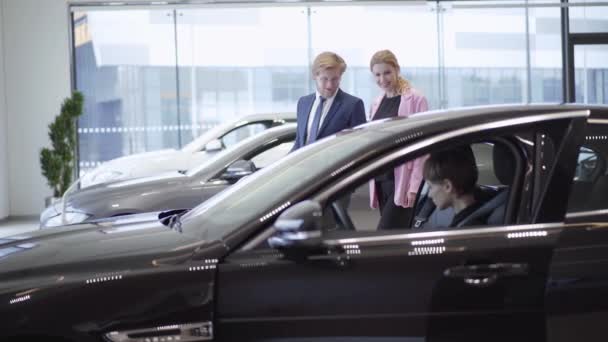 Selbstbewusste Frau mit kurzen Haaren sitzt im Auto vor lächelnden Geschäftsleuten und Verkäuferinnen und diskutiert über den Autokauf. Mann wählt Automobil für seine Frau in Autoshow — Stockvideo