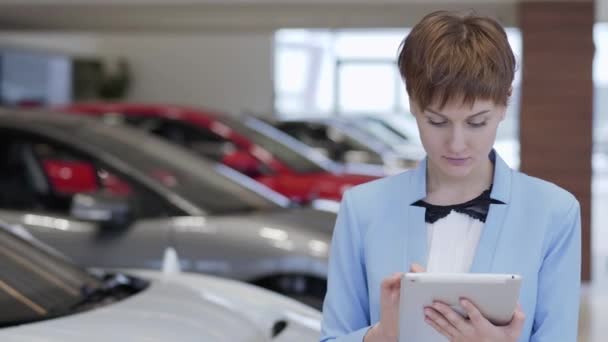 Porträt einer selbstbewussten hübschen Frau in formeller Kleidung mit ihrem Tablet steht vor verschwommenen Autos in der Autoschau. Verkäuferin hält Ordnung in modernem Autohaus — Stockvideo