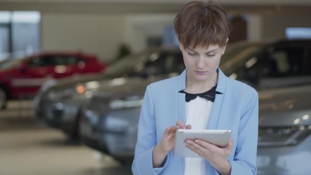 Portret van zeker mooie vrouw in formele slijtage met behulp van haar tablet staat voor wazig auto's in het Autosalon. Dame op zoek rond, vervolgens in de camera. Professionele verkoopster werkt in auto-winkel — Stockvideo