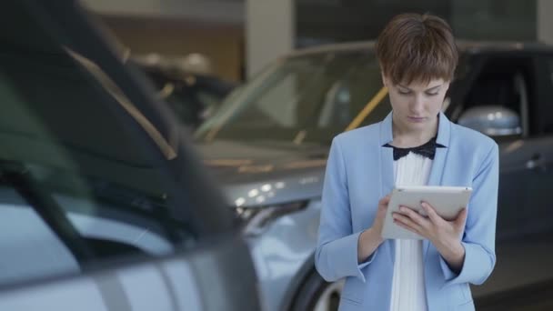 Retrato de mulher bonita confiante no desgaste formal usando seu tablet verificando carros no motor show. Senhora a examinar veículos. Vendedora profissional trabalha em loja de carros — Vídeo de Stock