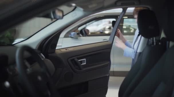 Mujer bonita joven en traje azul formal de desgaste abre la puerta del coche y se sienta dentro de ella en el asiento delantero del pasajero. Chica moderna elegir el automóvil para comprar, examinándolo en el espectáculo de motor. Concepto de compra de un — Vídeos de Stock