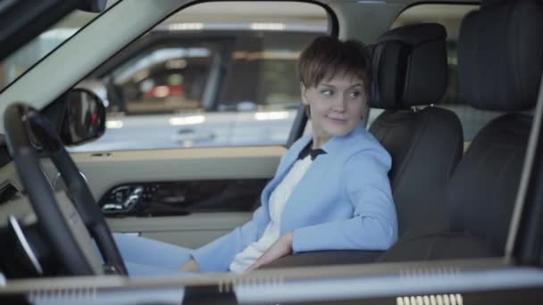 Young woman with short haircut in formal wear blue suit sitting inside a car in front passanger seat. Modern girl examining automobile interior, smiling. Concept of buying a vehicle in motor show — Stock Video