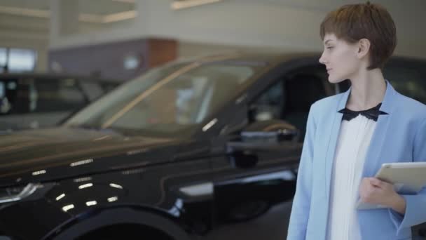 Linda vendedora com tablet mostra o carro no showroom. Vendedora de sucesso em pé na frente do veículo moderno demonstra mostra carro e olha para a câmera, sorrindo. Conceito de venda de automóveis — Vídeo de Stock