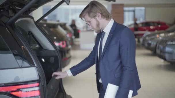 Homme d'affaires barbu choisissant la voiture dans le salon de l'automobile de près. Grand homme tenant comprimé regarde dans la manivelle ouverte et examine le véhicule. Homme sérieux choisissant l'automobile en salon de l'automobile. Concept d'achat de voiture — Video