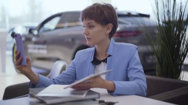 自信的女人, 穿着经典的蓝色正式西服短发, 坐在桌旁用手机在现代车展上使用前置摄像头交谈。后面的车模糊了。汽车陈列室的女售货员 — 图库视频影像