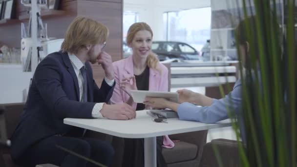 Unrecognized car dealer in a stylish suit shows information about their new car on the tablet to young successful couple. Car dealership. Motor car. — 图库视频影像