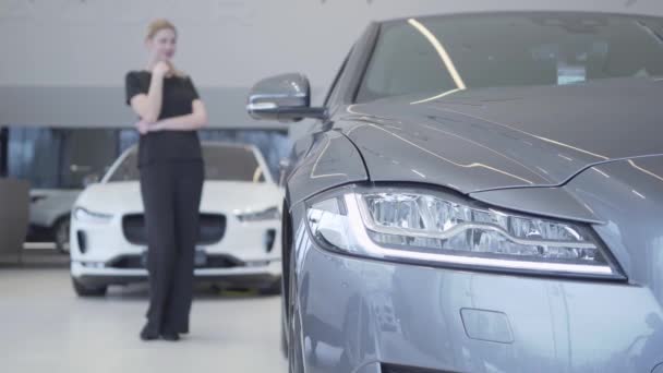 Wazig lachende vrouw figuur in de zwarte slijtage staande in Autosalon rondkijken. Koplamp van moderne zilveren auto op de voorgrond. Klant kiezen auto te kopen. Concept van het voertuig te kopen — Stockvideo