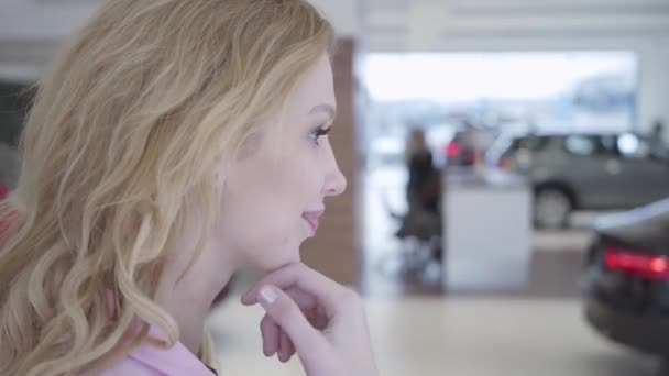 La cara de una chica de éxito reflexivo con el pelo rubio elegir un coche nuevo en el espectáculo de automóviles de élite. Sala de exposición de coches . — Vídeos de Stock