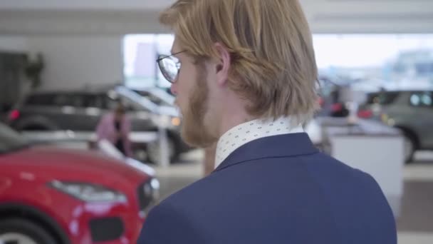 Close-up de um retrato pensativo de um homem de negócios em um terno e óculos tocando uma barba e escolhendo um carro em uma concessionária de carros. Sala de exposições do carro . — Vídeo de Stock