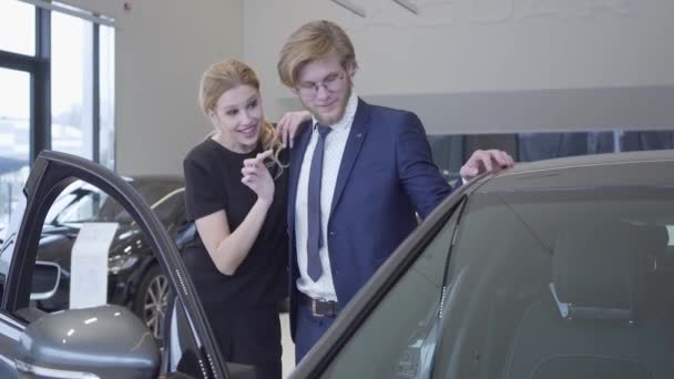 Joyeux homme et femme viennent d'acheter une voiture dans le salon de l'automobile moderne. Couple étreignant près de l'automobile avec porte ouverte dans un concessionnaire automobile. Homme d'affaires achetant un véhicule pour femme — Video