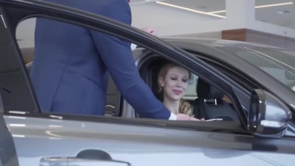 Le vendeur en costume d'affaires informe une jeune fille acheteuse d'une nouvelle voiture de luxe. Le concessionnaire donne à l'acheteur une tablette avec les caractéristiques du véhicule. Voiture automobile . — Video