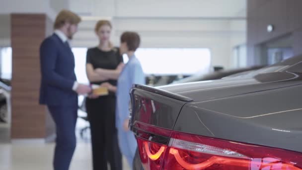 Verschwommene Figuren von Verkäuferinnen und zwei Kundinnen, die sich im Hintergrund moderner Autos auf Autosalons unterhalten. Frau wählt Fahrzeug für ihre Tochter. Konzept des Autokaufs — Stockvideo