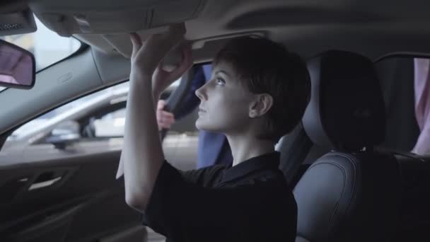 Jonge vrouw onderzoekt nieuwe auto zittend in de passagiersruimte van de nieuwe auto in auto showroom. De verkoper in een pak adviseert een succesvolle zakenvrouw. Automobielbedrijf. — Stockvideo