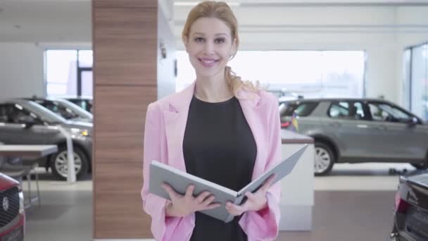 Retrato de um vendedor sorridente vestido em um terno de negócios que encontra visitantes em uma concessionária de carros. A menina vai encontrar compradores entre as fileiras com veículos de luxo caros. Concessionária de automóveis . — Vídeo de Stock