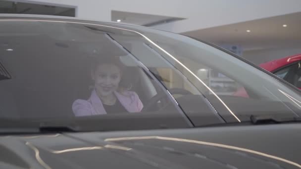 Jovencita bonita alegre en el compartimiento de pasajeros del nuevo auto en la sala de exposición del coche. La chica está llamando a un gerente de coche. concesionario de coches . — Vídeo de stock