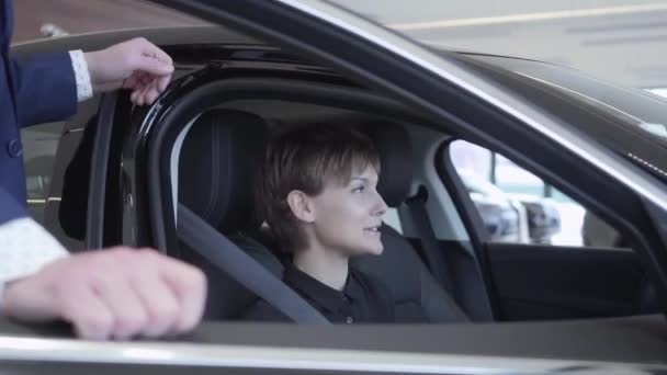 Mujer bonita con el pelo corto se sienta en el coche moderno hablando con vendedor irreconocible. Vendedor profesional en traje consultando al cliente. Concepto de compra de automóviles — Vídeos de Stock