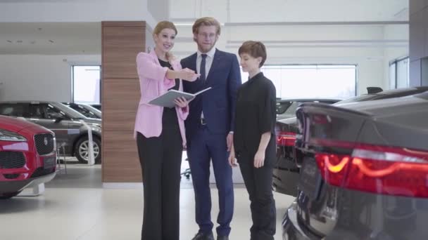 Saleswoman en veste rose consultation des clients couple sur la voiture. Homme d'affaires et sa femme choisissent le véhicule. Concept d'achat automobile, entreprise automobile — Video