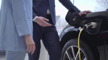 Kıdemli elektrik kablosunu elektrikli arabaya takıyor. Kadın müşteri diğer otomobiller üzerinde avantajlarını açıklayan elektrikli araba şarj yakınında takım elbise ayakta tanınmaz profesyonel satıcı.