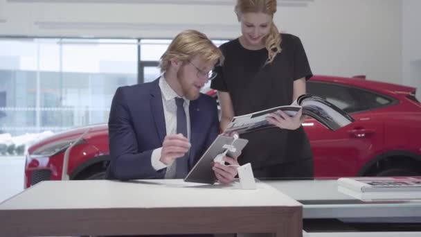 Une femme d'affaires prospère parcourt le catalogue avec des véhicules debout près d'un vendeur élégant en costume d'affaires qui s'assoit à une table dans un concessionnaire automobile. Voiture automobile . — Video