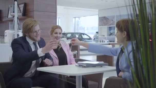 Saleswoman dans un costume élégant transmettant les clés de la nouvelle voiture heureux couple d'affaires réussi. Un barbu joyeux en costume d'affaires prend joyeusement les clés de son nouveau véhicule. Salle d'exposition de voiture — Video