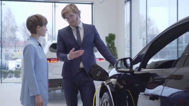 Vendeur professionnel confiant raconte à une jolie jeune femme d'affaires dans un costume élégant les détails et les caractéristiques d'une nouvelle voiture électrique debout au véhicule de charge dans le concessionnaire automobile . — Video