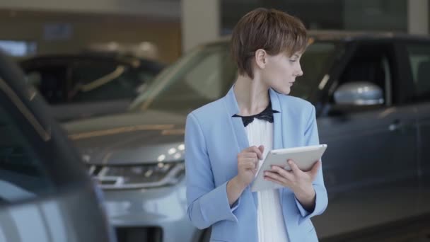 Retrato de mulher bonita confiante em um terno elegante usando seu tablet verificando carros no motor show. Senhora a examinar veículos. Vendedora profissional trabalha em loja de carros — Vídeo de Stock