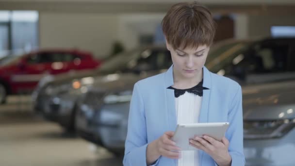 Portré magabiztos aranyos nő hivatalos viselet használ vele tabletta áll előtt homályos autók Autószalonon. Hivatásos elárusítónő működik autókereskedés — Stock videók