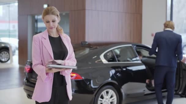 Portrait of pleasant cute girl in pink jacket with a big book about cars in front of couple choosing vehicle. Man opens the door and woman sits inside. Concept of buying automobile, auto business