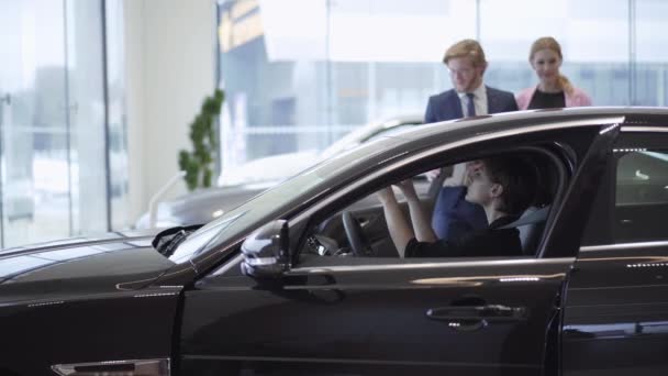 Jovem bonita feliz no compartimento de passageiros do carro novo. Mulher bem sucedida examina novo automóvel. Jovem casal de sucesso escolhendo seus próprios passeios de carro em segundo plano. Concessionária de automóveis . — Vídeo de Stock