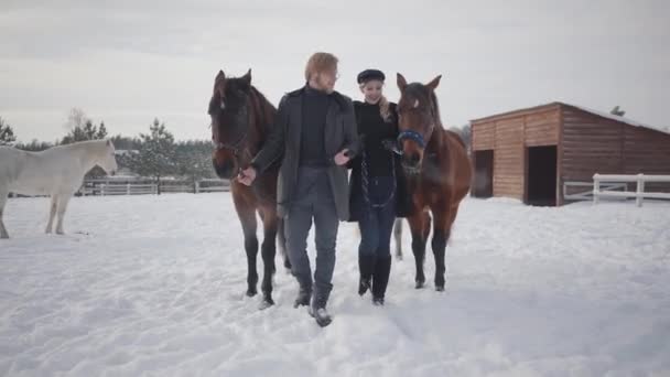 Ein lächelndes Paar, das im Winter auf einer Ranch im Freien mit Pferden spaziert. Mann und Frau an der Spitze der Pferde. Pferde und Ponys folgen dem Paar. — Stockvideo