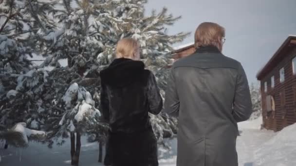 Hombre y mujer en abrigos calientes caminando en el patio de nieve cerca de casas de madera hacia la figura masculina con caballo blanco. Familia joven descansa al aire libre en invierno — Vídeos de Stock