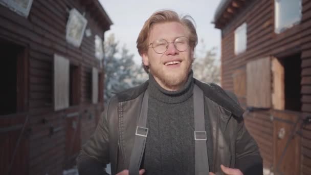 Retrato de un guapo barbudo con gafas vestidas con un suéter cálido y una chaqueta abierta. El tipo sonríe, levanta tirantes y mira a su alrededor . — Vídeos de Stock