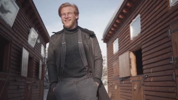 Portret van een gelukkig bebaarde man in glazen gekleed in een warme trui en wijdopen jas. Jongen glimlacht en kijkt rond. — Stockvideo