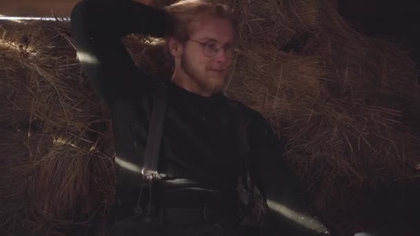 Retrato de un guapo barbudo con gafas sentado en un heno en un establo. El tipo sacude el heno de su suéter . — Vídeo de stock