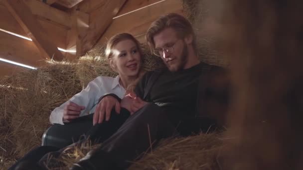 Retrato de casal muito jovem deitado no feno no celeiro frio de inverno. Jovem agricultor descansando após o dia de trabalho com sua esposa aquece as mãos. Mulher brinca com palha — Vídeo de Stock