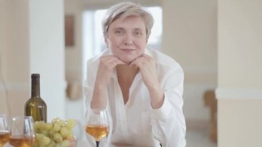 Kısa saç kesimi, kamera kapalı hoş gülümseme ile seyir ile beyaz bluz çok şık üst düzey kadın portresi. Şarap ve meyve mutfak tabloyla yakın duran Olgun kadın.