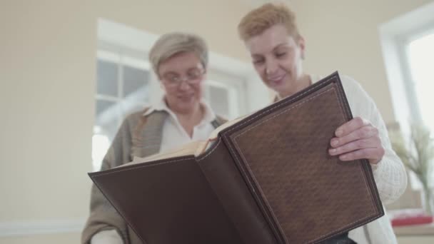 Zwei ältere Frauen halten ein großes Fotoalbum aus Leder in der Hand und sehen sich Fotos an. Reife Frauen mittleren Alters kommunizieren im Chat und verbringen Zeit zu Hause — Stockvideo