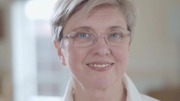 Close up retrato de mulher sênior com óculos olhando na câmera com sorriso agradável dentro de casa . — Vídeo de Stock
