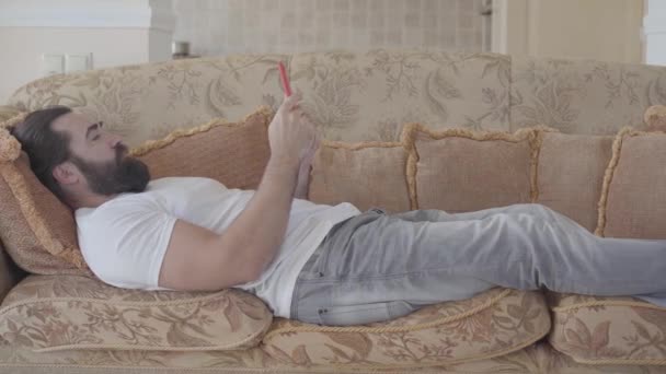 Homem com barba no sofá bege macio no meio do dia relaxando e digitando sms ou procurando algo na internet — Vídeo de Stock
